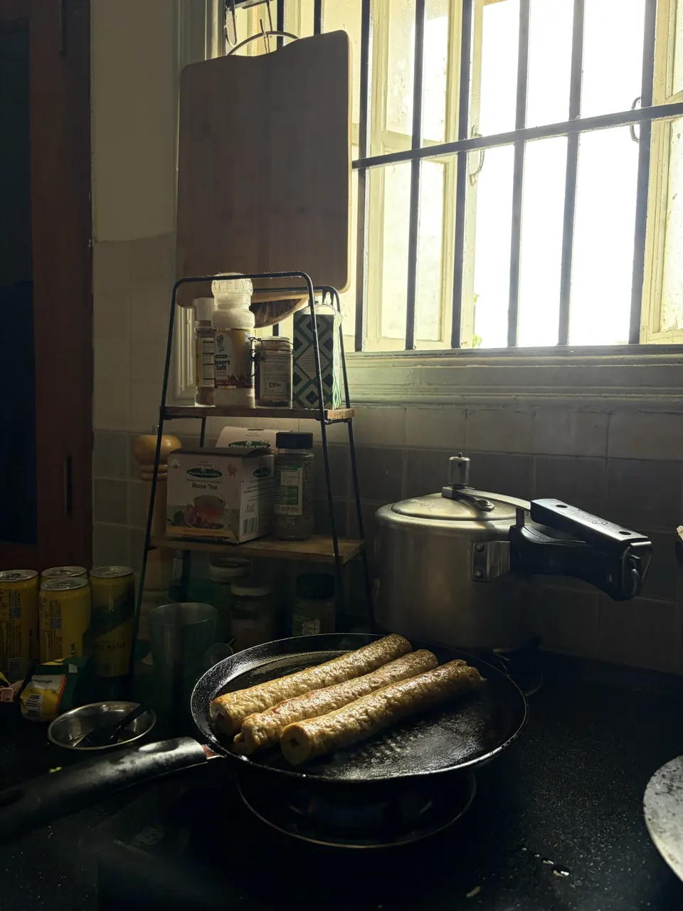 Rolled flatbreads crisping on an iron tawa in a dim kitchen lit by a barred window