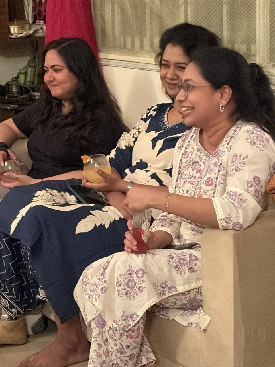 Three women in printed kurtas laugh together on a sofa holding drinks