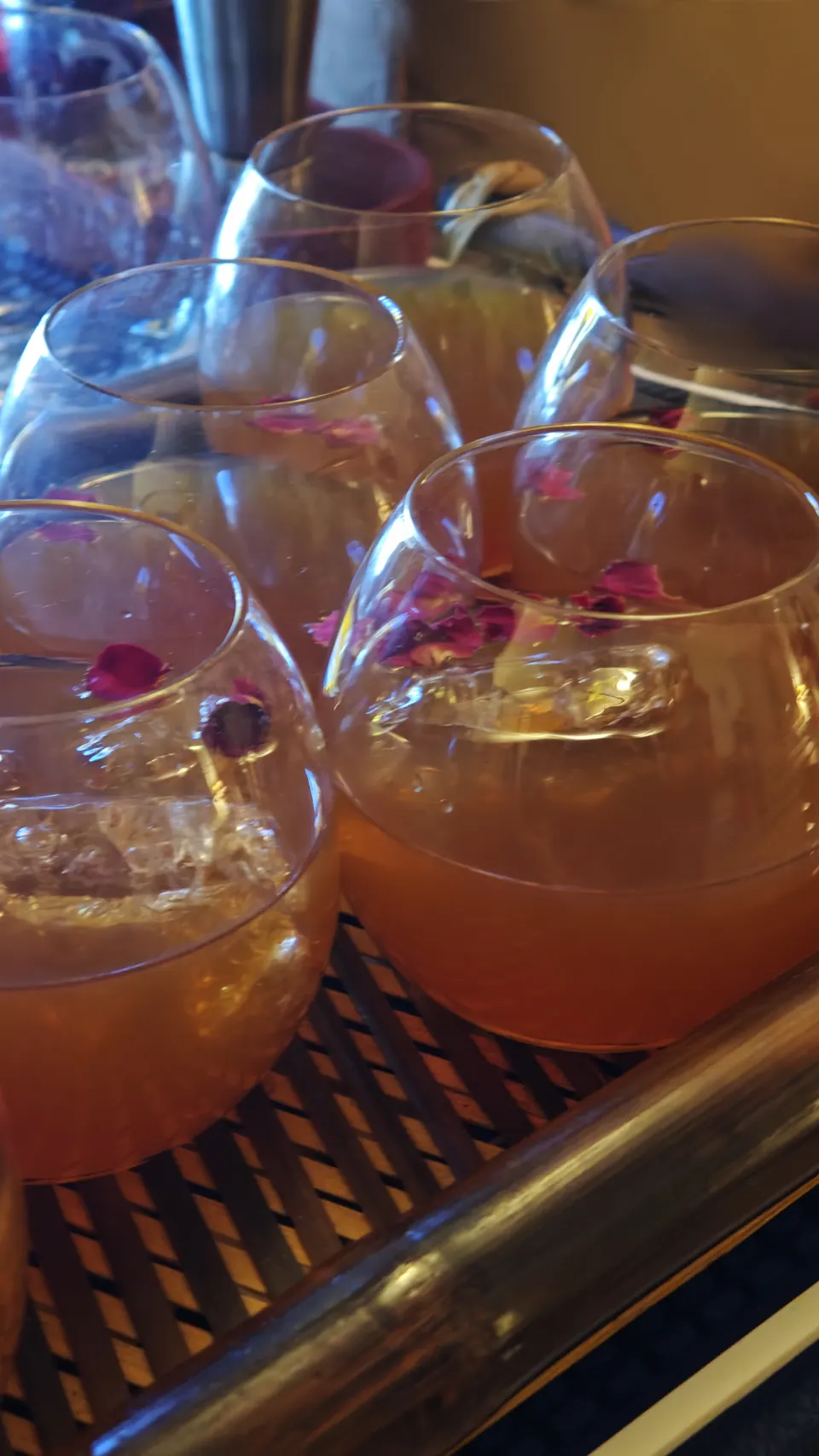 A tray of whisky cocktails in round glasses, each topped with dried rose petals