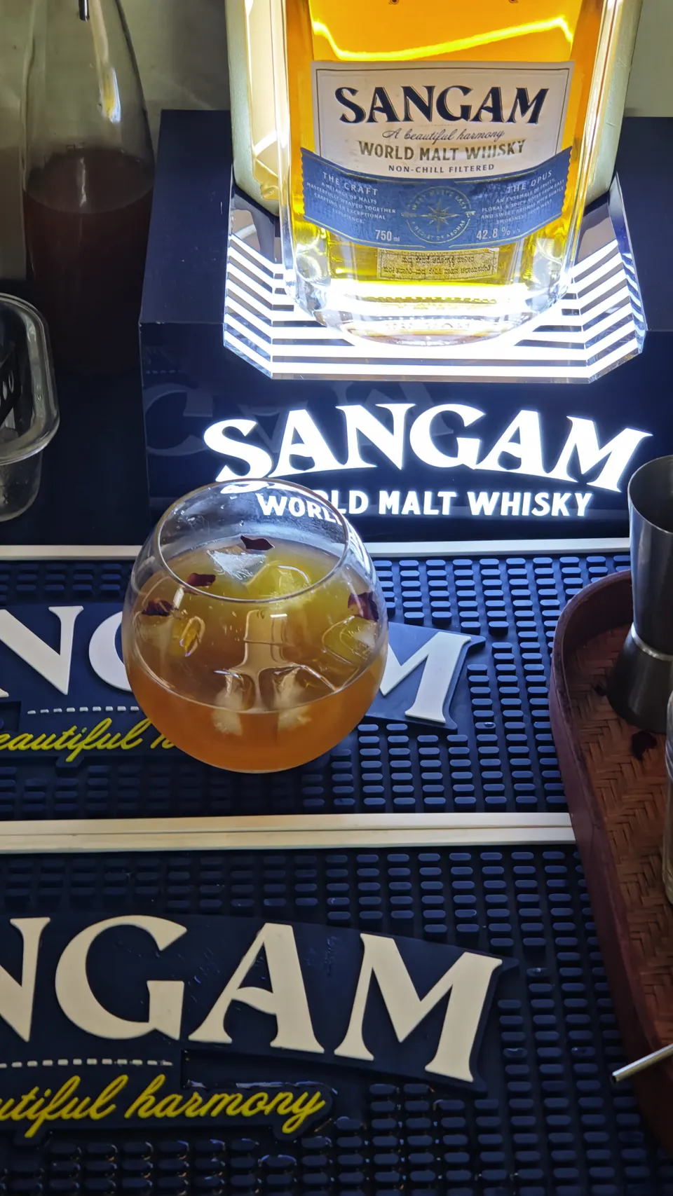 Rose-petal whisky cocktail on a navy Sangam bar mat beneath a lit whisky bottle display