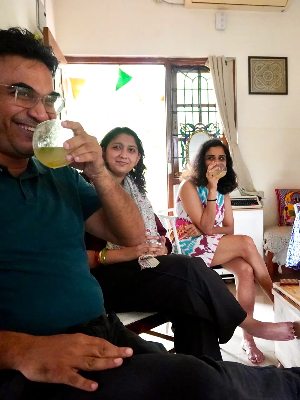 A grinning guest sips a pale green drink while two women seated by a bright doorway look on