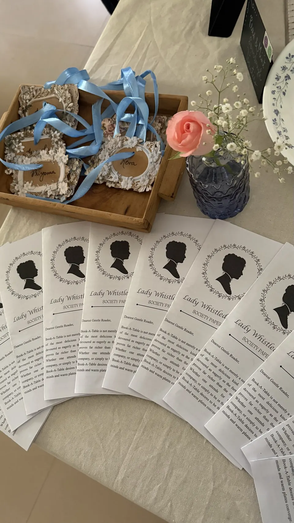 Fanned Lady Whistledown's Society Papers beside ornate floral name tags with blue ribbons and a pink rose