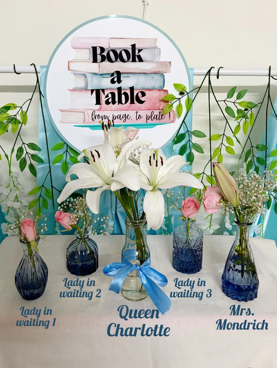 Five blue glass vases of lilies and roses before the Book a Table sign, cast as Queen Charlotte's court