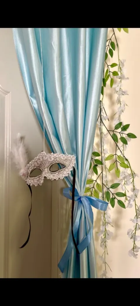 Silver lace masquerade mask hanging against a powder-blue satin curtain tied with ribbon and white wisteria