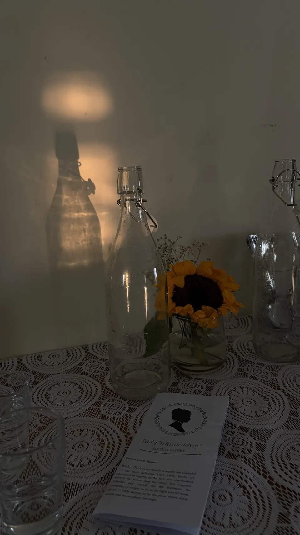 Glass swing-top bottles and a sunflower on lace casting golden shadows, with a Whistledown paper in front