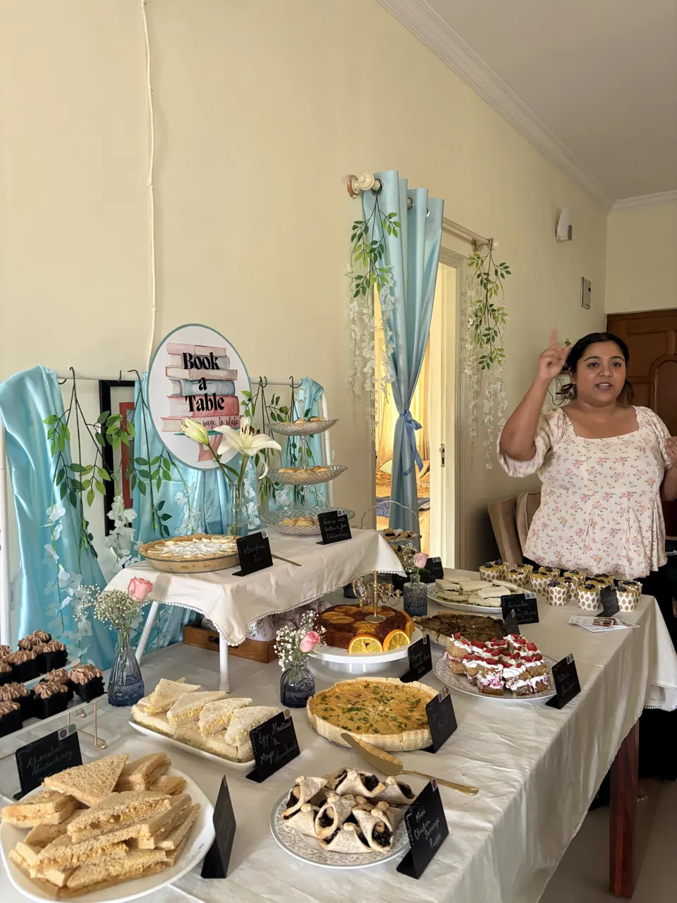 Host in a floral dress introducing the full afternoon-tea spread of quiches, sandwiches, cakes and muffins