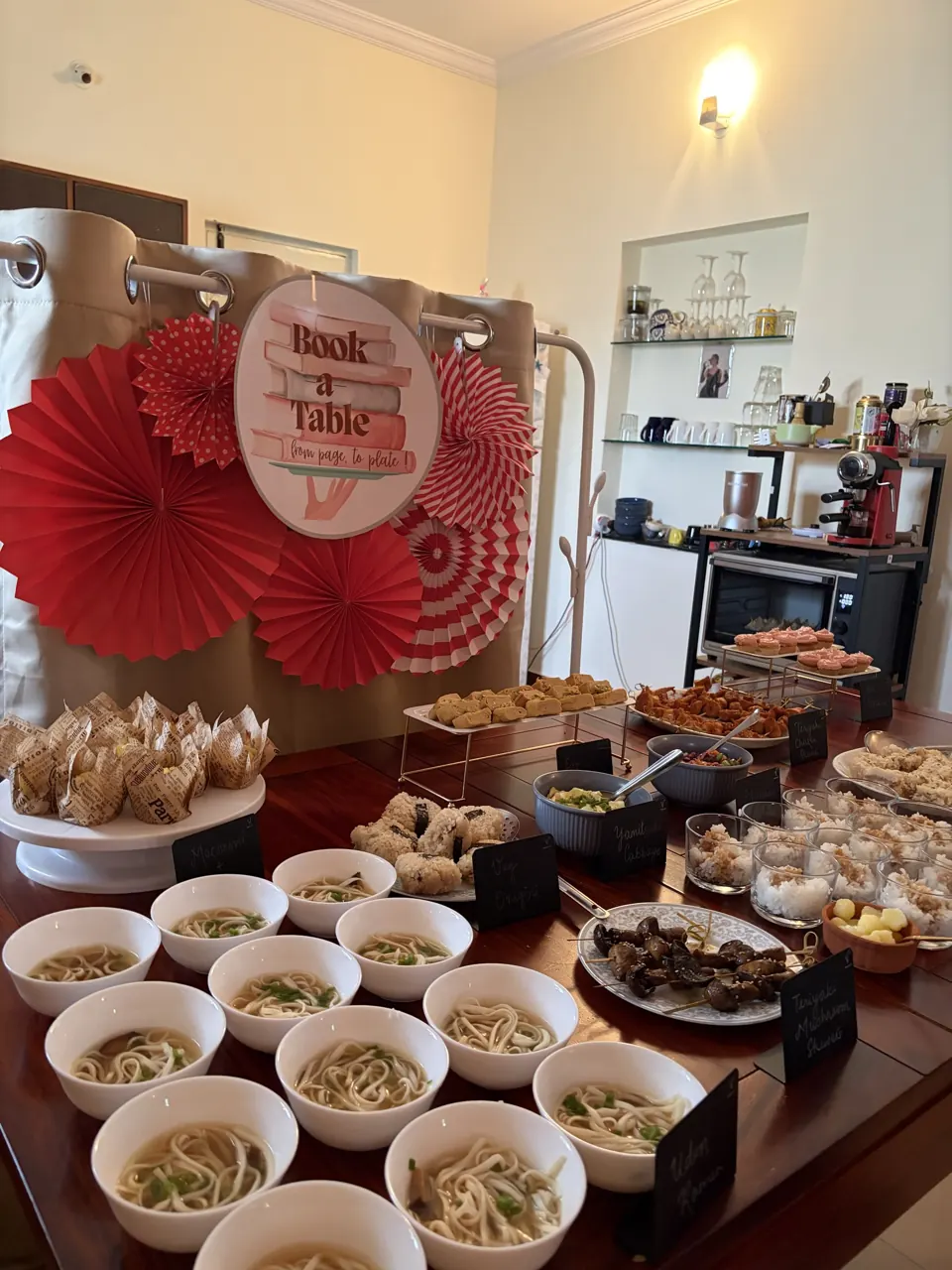 Japanese-inspired buffet of udon bowls, onigiri, skewers and cupcakes with chalkboard tags beside the Book-A-Table sign