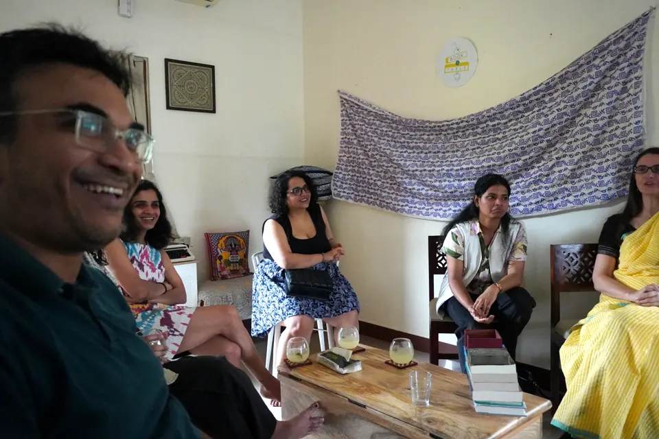 Laughing guest in foreground with others seated around a coffee table stacked with books