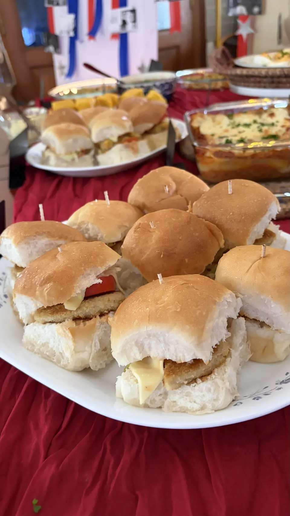 Platter of cheese and tomato sliders on a red tablecloth, baked casserole behind